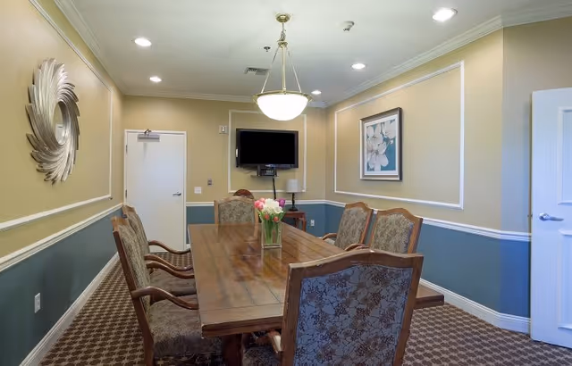 A well-lit dining room with a long wooden table surrounded by six upholstered chairs. A vase with pink and white tulips is placed in the center of the table. The walls are painted beige and blue with white trim, decorated with a round mirror and a framed floral painting. A flat-screen TV is mounted on the far wall above a small table with a lamp. The room has a patterned carpet and a ceiling light fixture.