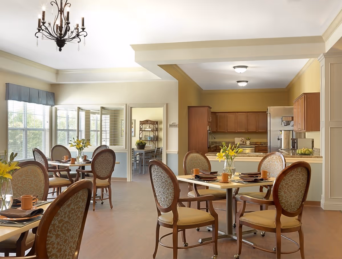 A bright dining area in a senior living facility with round tables set with brown napkins, orange cups, and vases of yellow flowers. The room has large windows with blinds, wooden chairs with patterned upholstery, and a chandelier hanging from the ceiling. In the background, there is a kitchen area with wooden cabinets and stainless steel appliances.