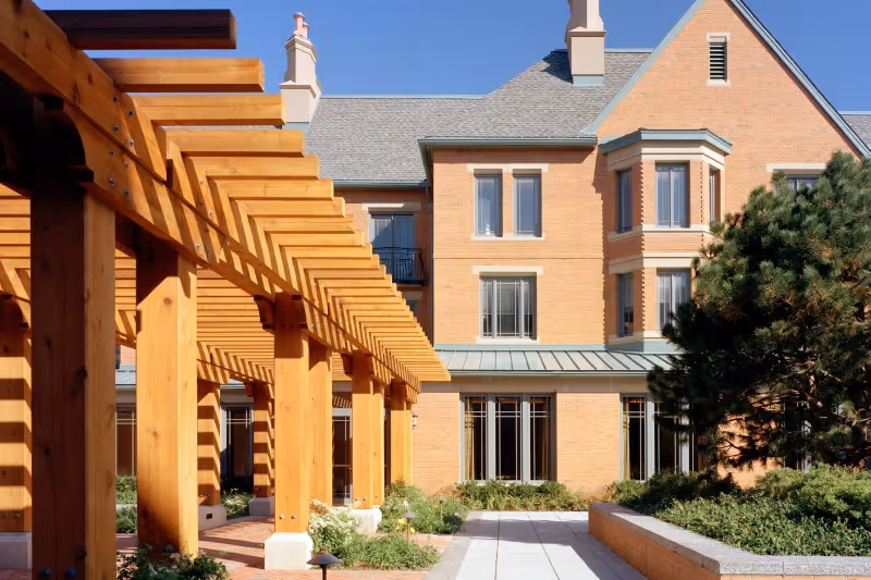 Exterior view of a brick building with large windows and a gray roof under a clear blue sky. In the foreground, there is a wooden pergola casting shadows on a paved walkway surrounded by greenery and shrubs.