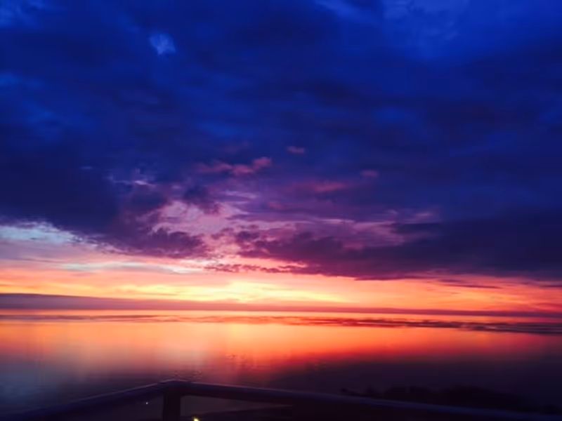 A vibrant sunset over a calm body of water with a railing in the foreground. The sky is filled with deep blue, purple, pink, and orange hues reflecting on the water surface.