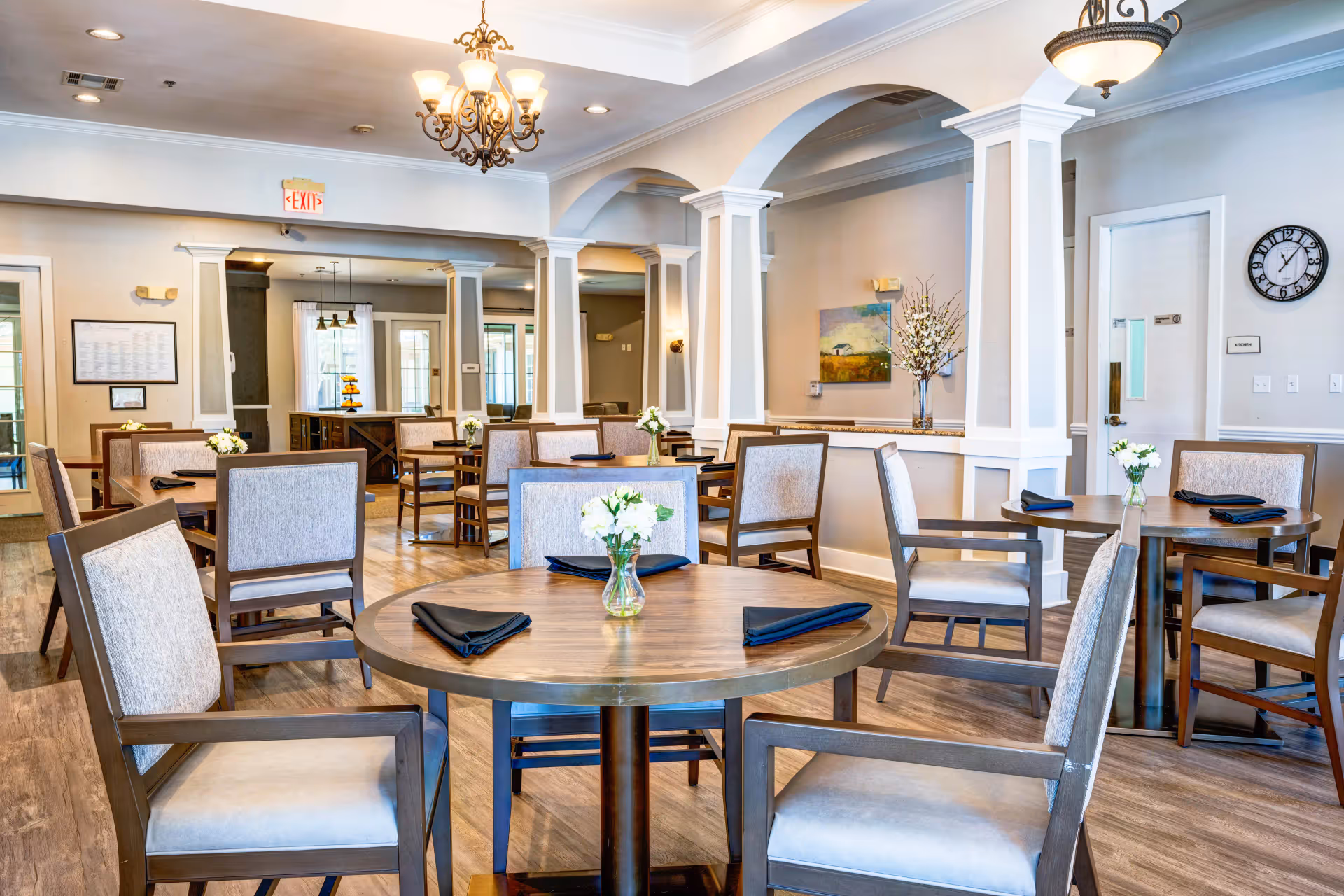 A bright and spacious dining room in an assisted living facility with multiple round wooden tables, each set with black napkins and small vases of white flowers. The room features light-colored walls, decorative columns, chandeliers, and a clock on the wall near a door labeled 'Kitchen'.