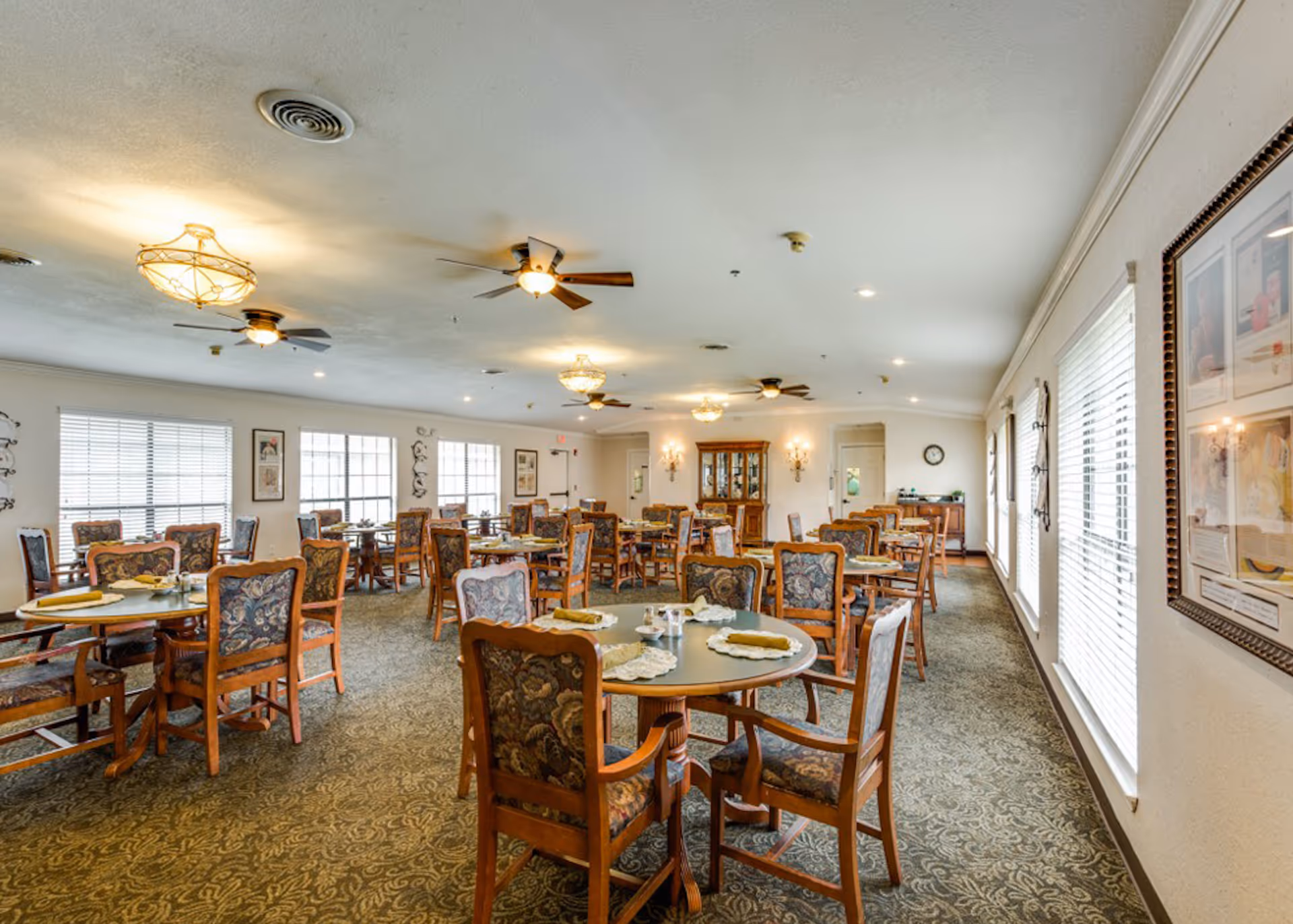 A spacious dining room with multiple round tables, each set with placemats, napkins, and salt and pepper shakers. The chairs have wooden frames and patterned upholstery. The room has large windows with blinds, carpeted floors, ceiling fans, and decorative light fixtures. There are framed pictures on the walls and a wooden cabinet at the far end of the room.