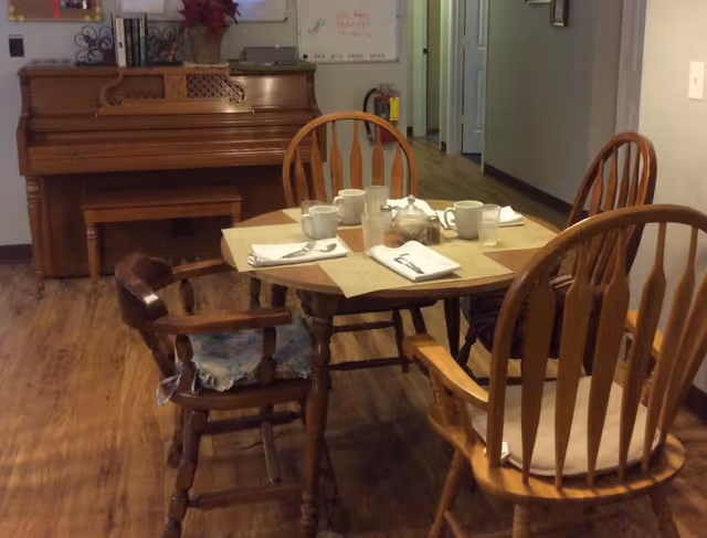 Wooden dining table with four chairs and place settings in a common area, with a piano and hallway visible in the background.