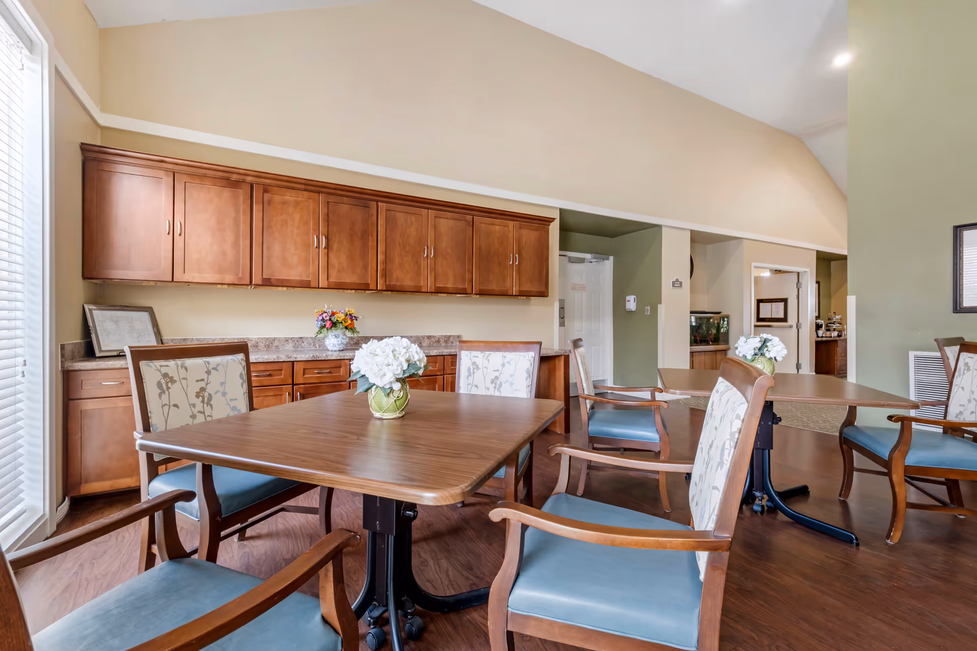 A bright and spacious common area with wooden tables and chairs featuring blue cushions and floral-patterned backs. The room has wooden cabinets along one wall, a vase with white flowers on each table, and a large window letting in natural light. The walls are painted in neutral tones with a high ceiling.