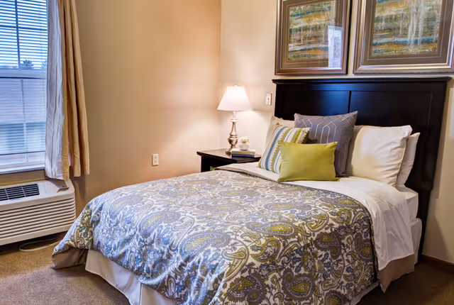 A neatly made bed with a patterned blue and green bedspread, several pillows including a green accent pillow, and a dark wooden headboard. Next to the bed is a nightstand with a lamp, a small plant, and a book. A window with beige curtains and an air conditioning unit is visible on the left side of the room. Two framed artworks hang above the headboard.