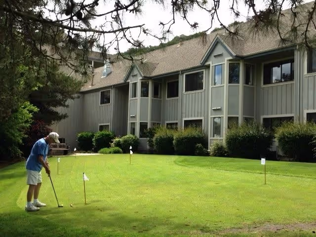 An elderly man wearing a blue shirt, white shorts, and a white cap is playing golf on a putting green outside a two-story residential building with gray siding and multiple windows. The green has several small flags indicating holes, and there are bushes and trees surrounding the area.