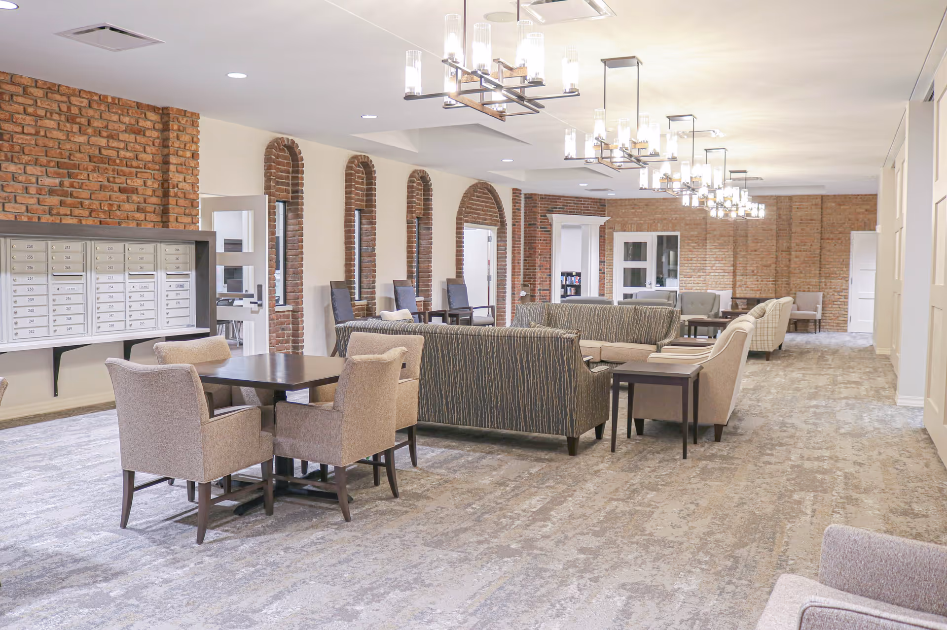 A spacious, well-lit common area with multiple seating arrangements including armchairs and sofas. The room features exposed brick walls with arched windows, modern ceiling lights, and a set of mailboxes mounted on one wall. The floor is carpeted with a neutral pattern, and the overall atmosphere is welcoming and comfortable.