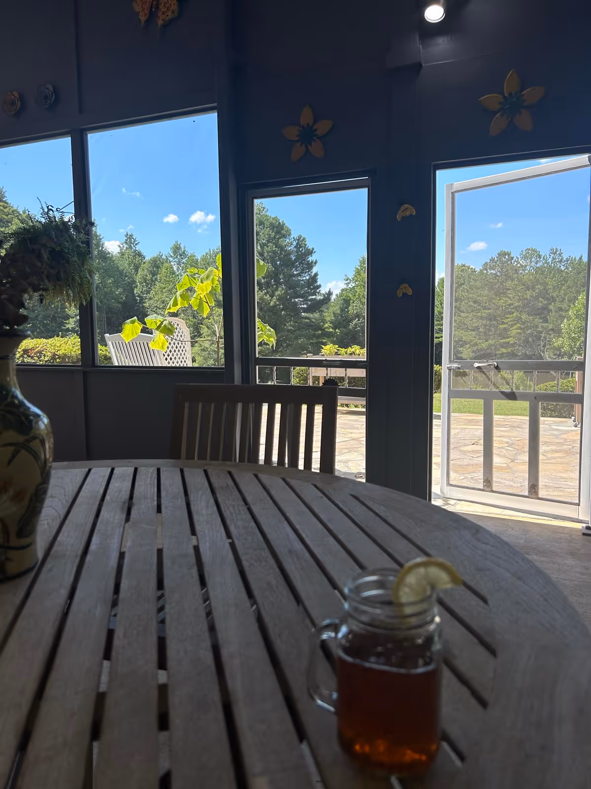 Wooden dining table with a mason jar of iced tea and chairs in a sunroom opening onto a sunny patio and trees outside.