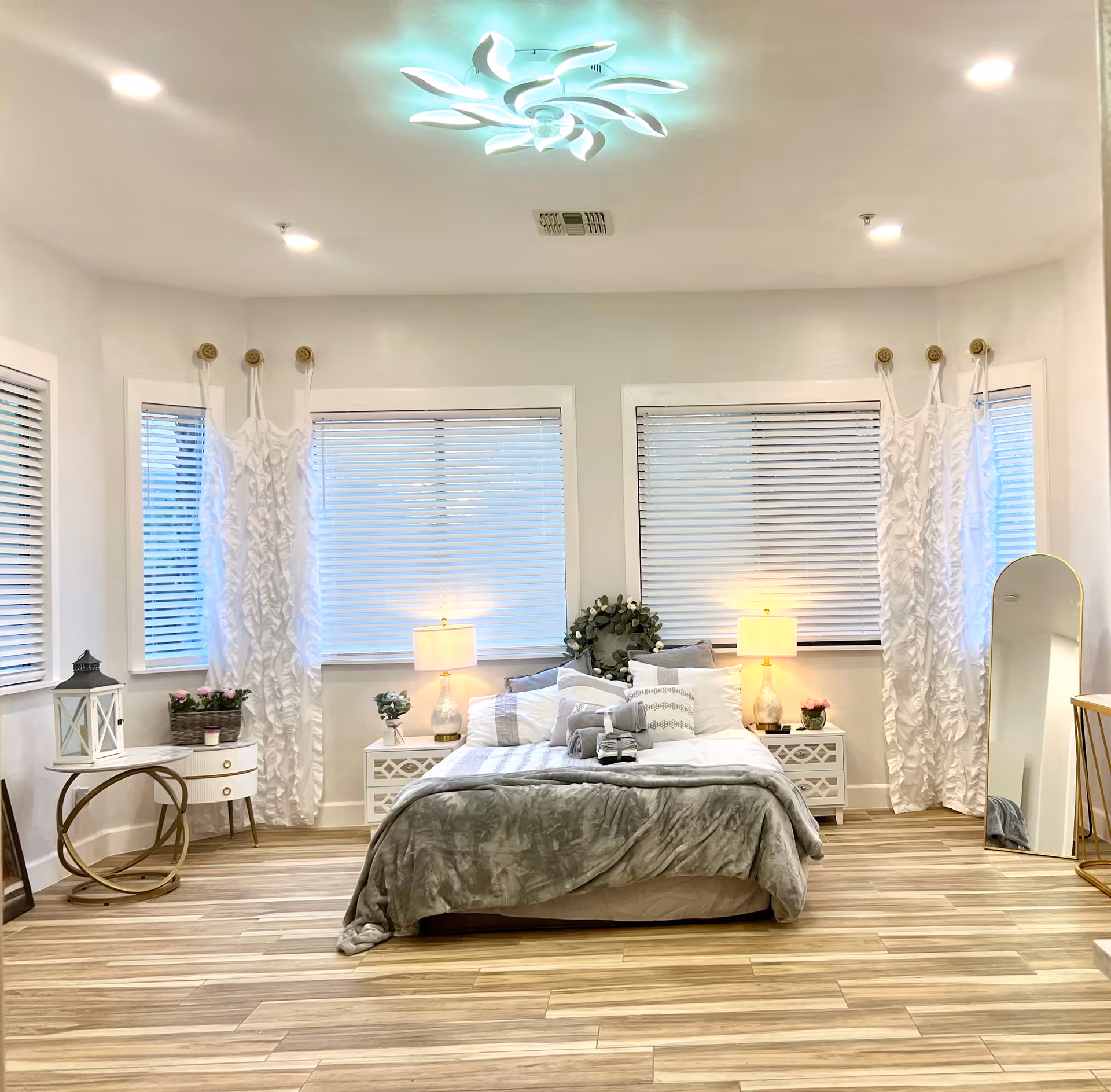 Bright modern bedroom with a centered bed, bedside tables and lamps, three large windows with white blinds and ruffled curtains, a floor mirror, and wood-look flooring.
