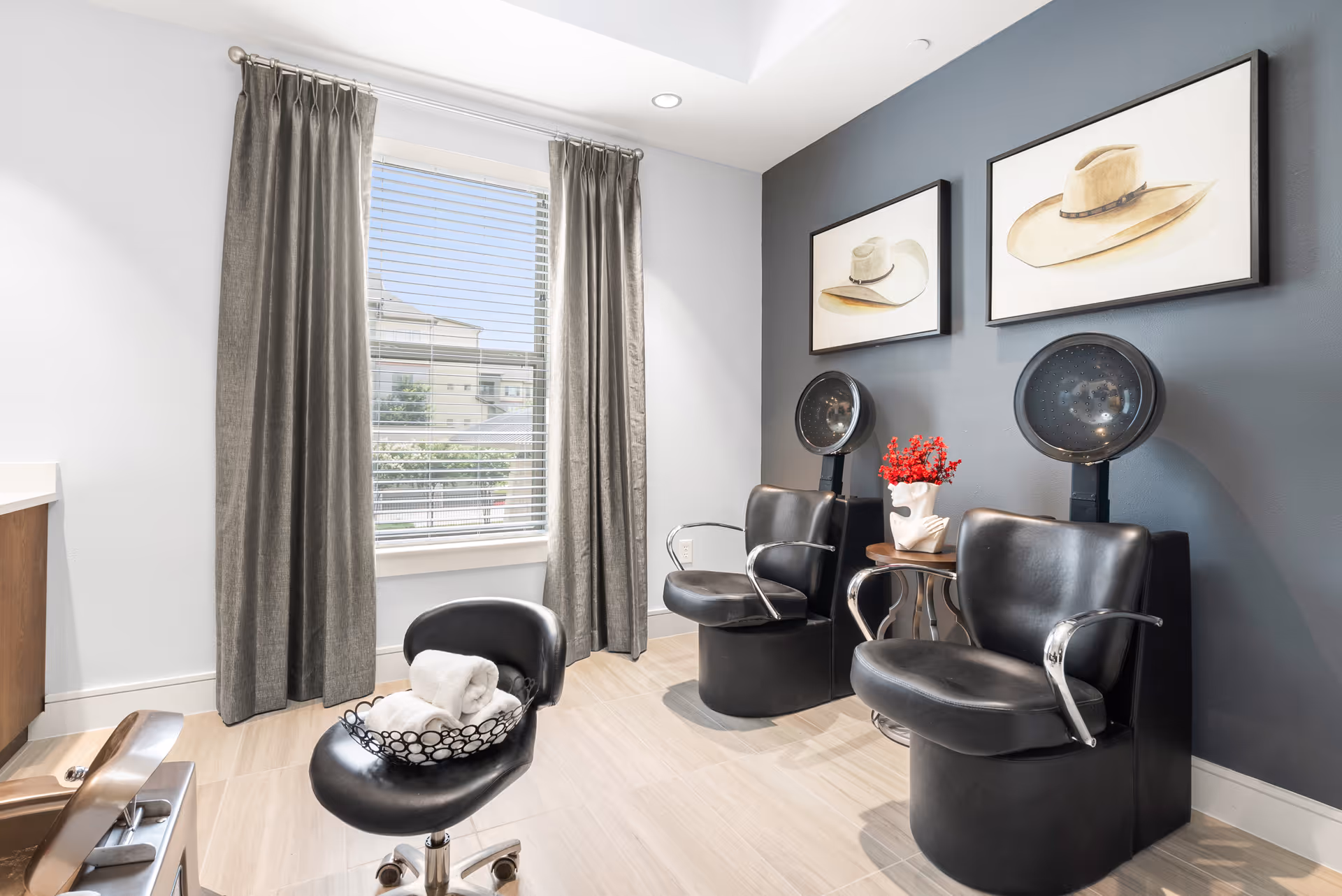 A bright salon-style interior with two black hooded dryer chairs, a rolling stool with towels, framed hat artwork, and a window with gray curtains.