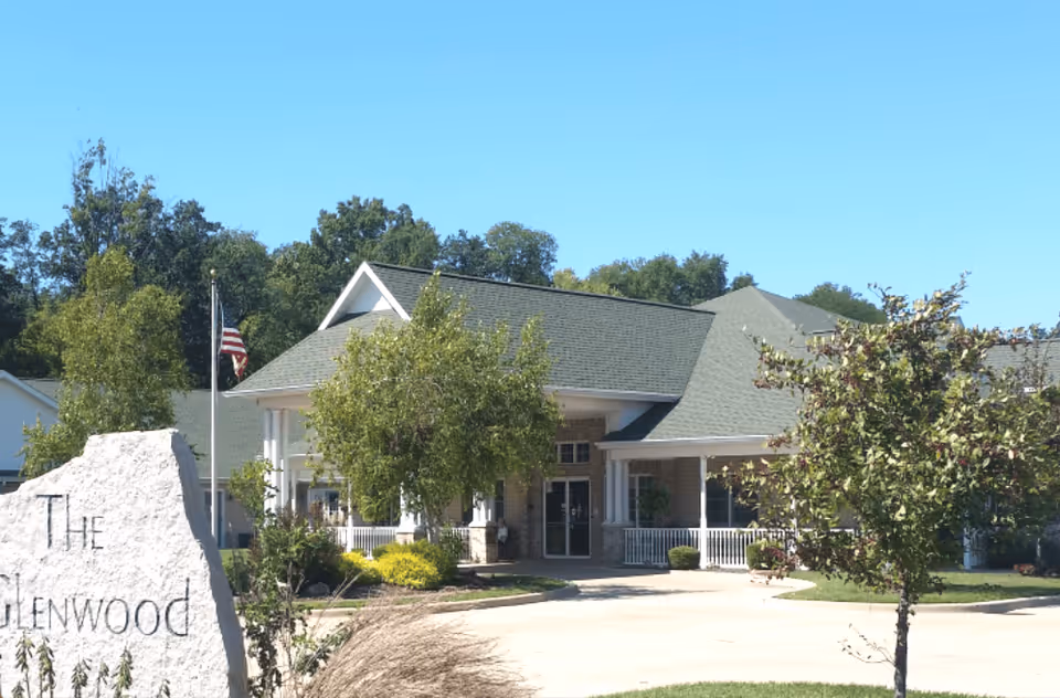 Exterior view of The Glenwood Supportive Living of Greenville building with a large stone sign in the foreground, trees, and a clear blue sky.