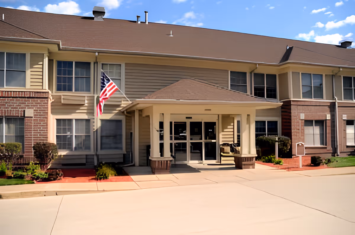 Exterior view of a senior living facility building with a covered entrance, beige and brick facade, multiple windows, and an American flag mounted near the entrance under a blue sky with some clouds.