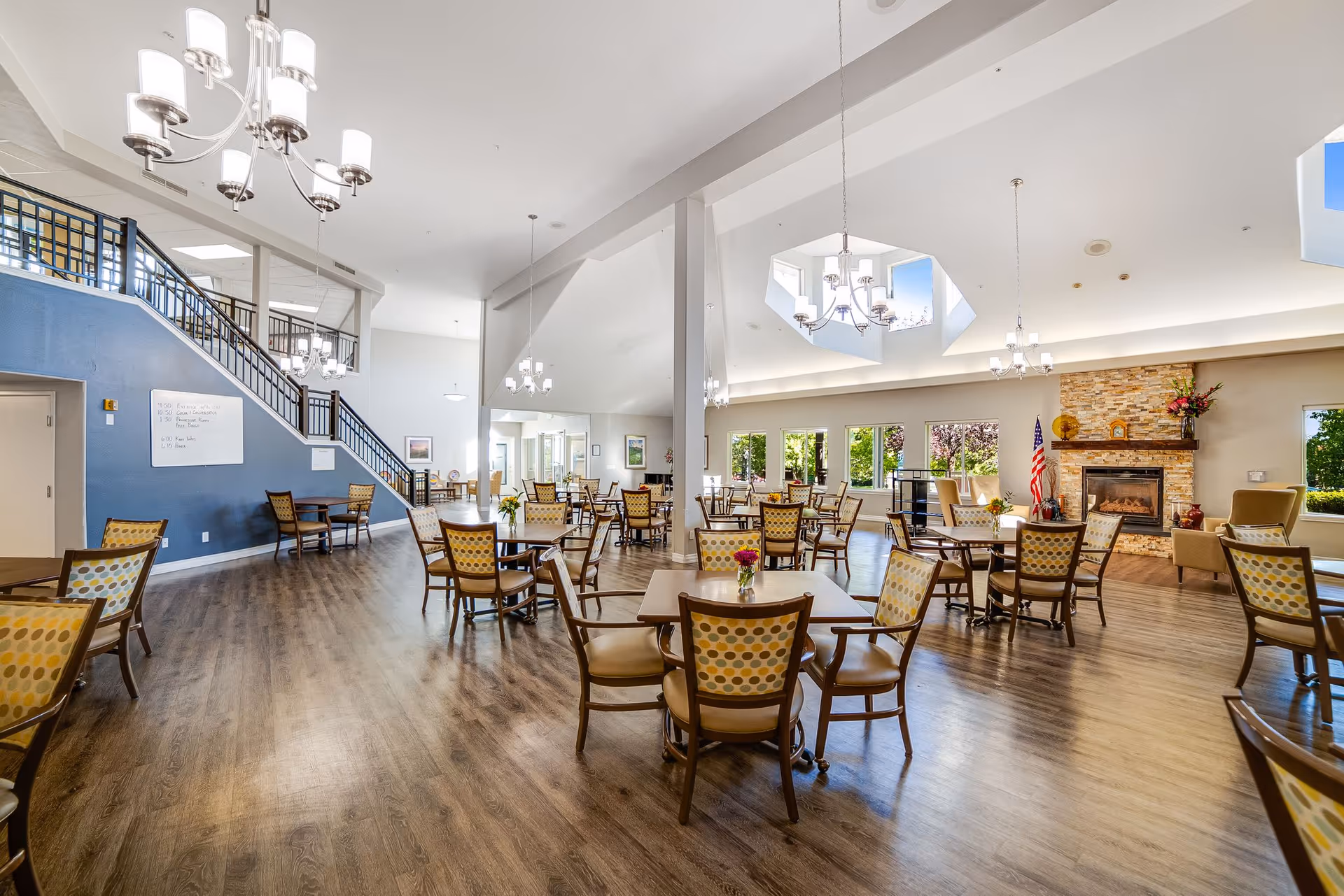 Spacious dining room with multiple wooden tables and chairs featuring patterned upholstery. The room has large windows letting in natural light, a stone fireplace with an American flag beside it, and modern chandeliers hanging from a high ceiling with skylights. A staircase with a black railing is visible on the left side, and there are flowers on some tables.