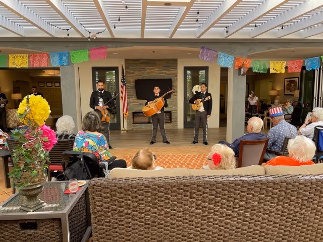 A group of elderly people seated in a common area watching a live musical performance by three musicians playing string instruments. The room is decorated with colorful paper banners hanging from the ceiling. The setting appears to be a senior living facility with comfortable seating and a fireplace in the background.