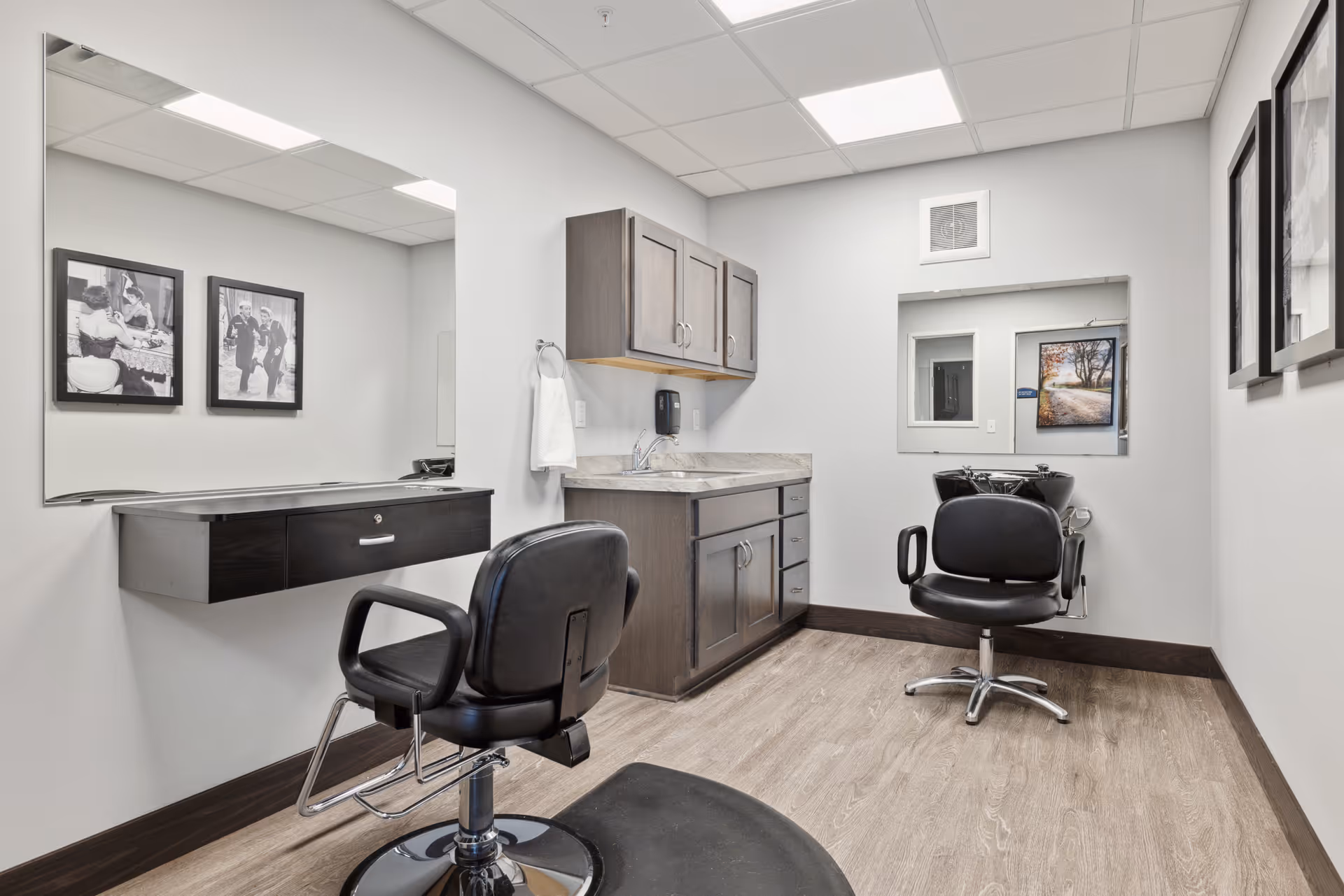 Interior of a senior living facility hair salon with two black salon chairs, a large mirror on the left wall, dark wood cabinets with a countertop and sink, and framed black and white photos on the walls.
