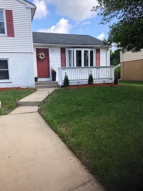 Front exterior of a house with a small white porch, red door and shutters, and a concrete walkway across a green lawn.