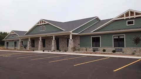 Exterior view of a single-story assisted living facility building with green siding, stone accents, and a gray roof. The building has a covered entrance supported by stone pillars and a large empty parking lot in front.