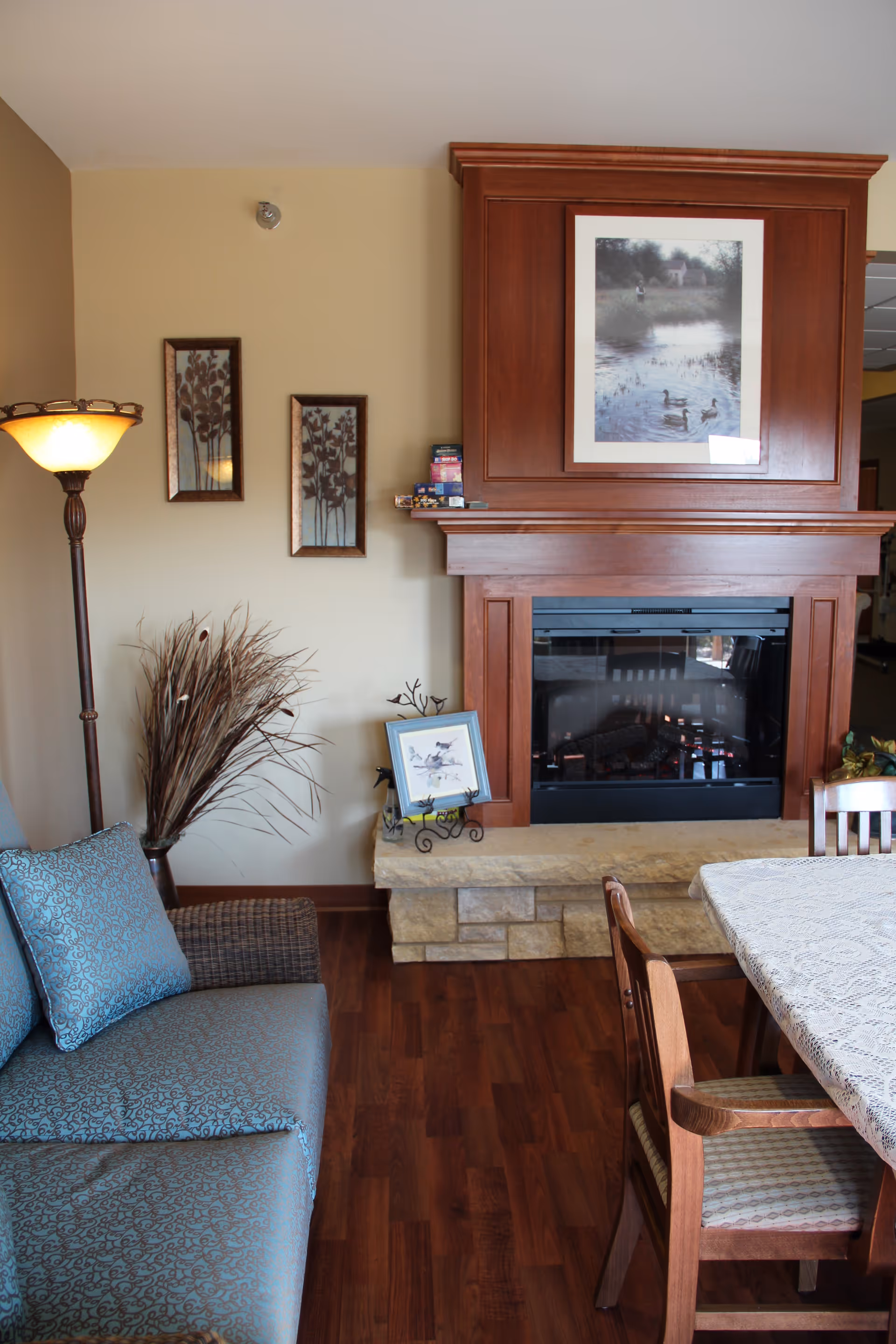Cozy living area with a wooden fireplace mantle, framed artwork, a sofa, floor lamp and a dining table.