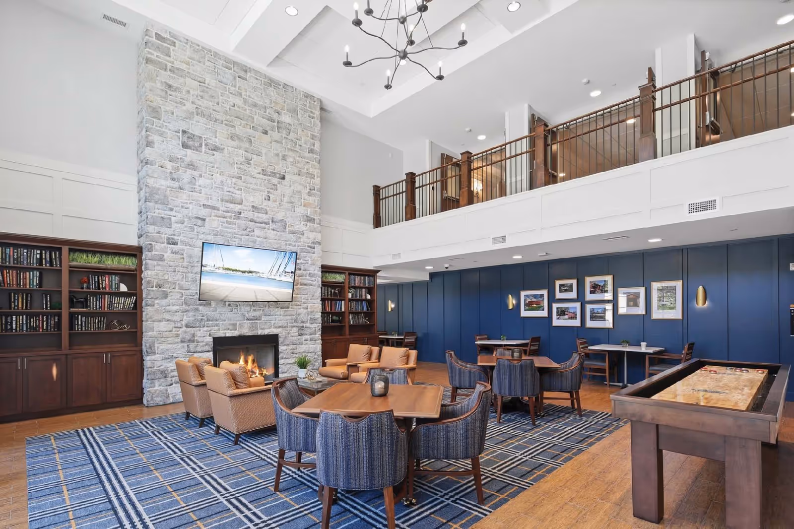 A spacious and well-lit common area with a high ceiling featuring a modern chandelier. The room has a large stone fireplace with a flat-screen TV mounted above it, flanked by built-in wooden bookshelves filled with books and decorative items. There are several seating arrangements including tan armchairs around the fireplace and blue upholstered chairs around wooden tables on a blue patterned carpet. The walls are painted blue with framed pictures, and there is a shuffleboard table on the right side of the room. A balcony with wooden railings overlooks the area.