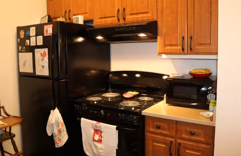 A kitchen with wooden cabinets, a black refrigerator with children's drawings attached, a black electric stove with coil burners, a microwave on the countertop, and a bowl of bananas. There are towels hanging on the refrigerator and stove handles.