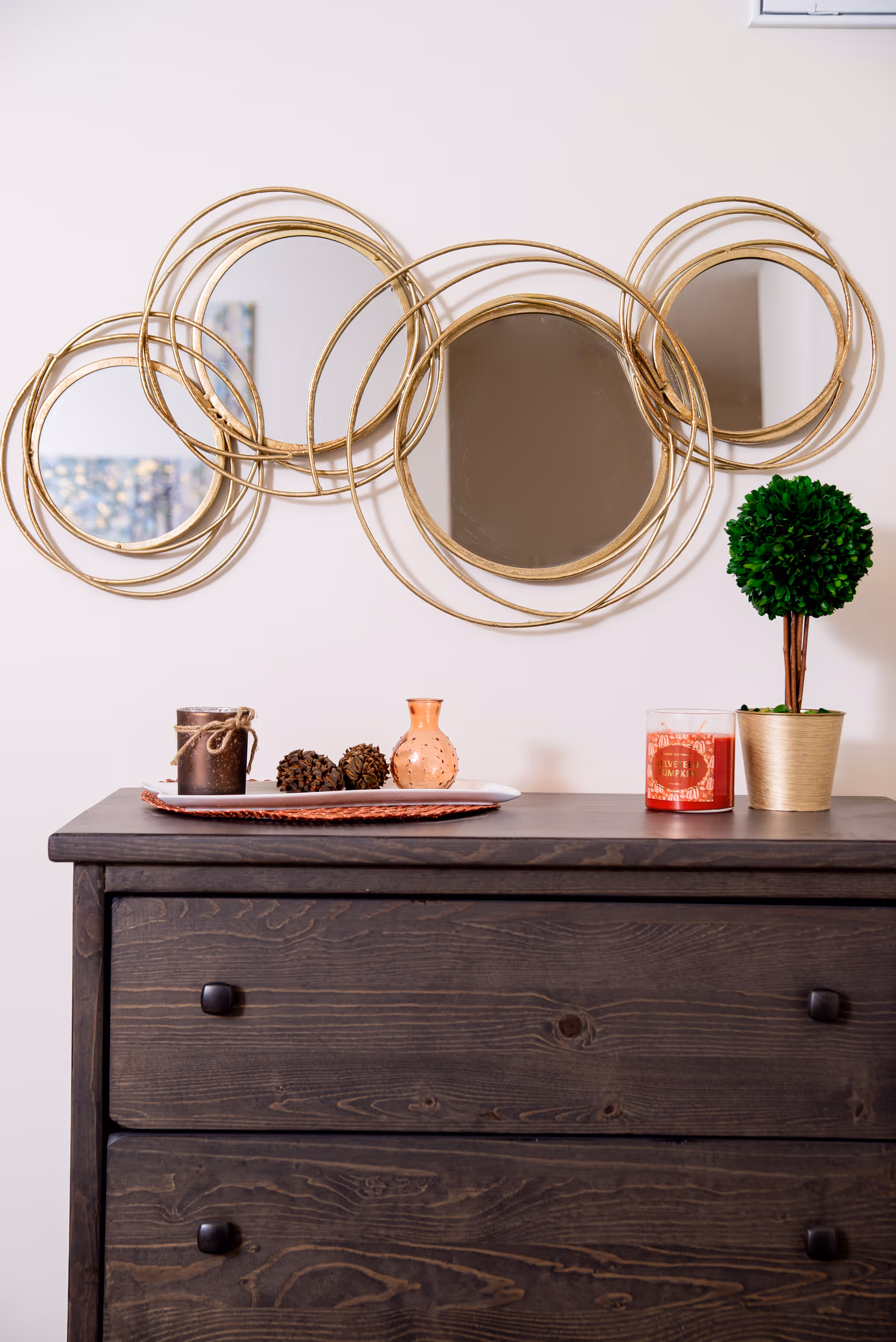 A dark wooden dresser with two drawers, decorated on top with a small potted artificial plant, a red candle, a small orange vase, pine cones, and a brown candle. Above the dresser is a wall decoration consisting of five round mirrors framed by overlapping gold rings.