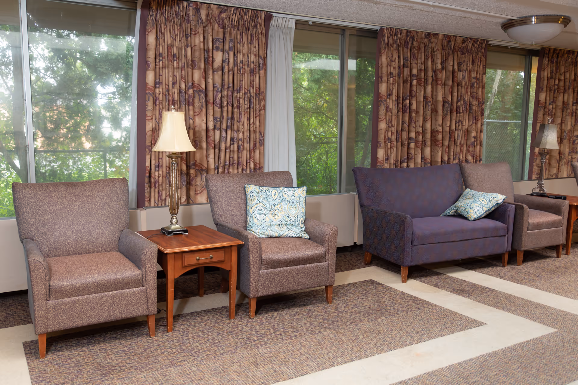 A seating area with two brown armchairs and a blue loveseat, each with decorative pillows. Between the chairs are wooden side tables with lamps. Large windows with patterned curtains reveal green trees outside.