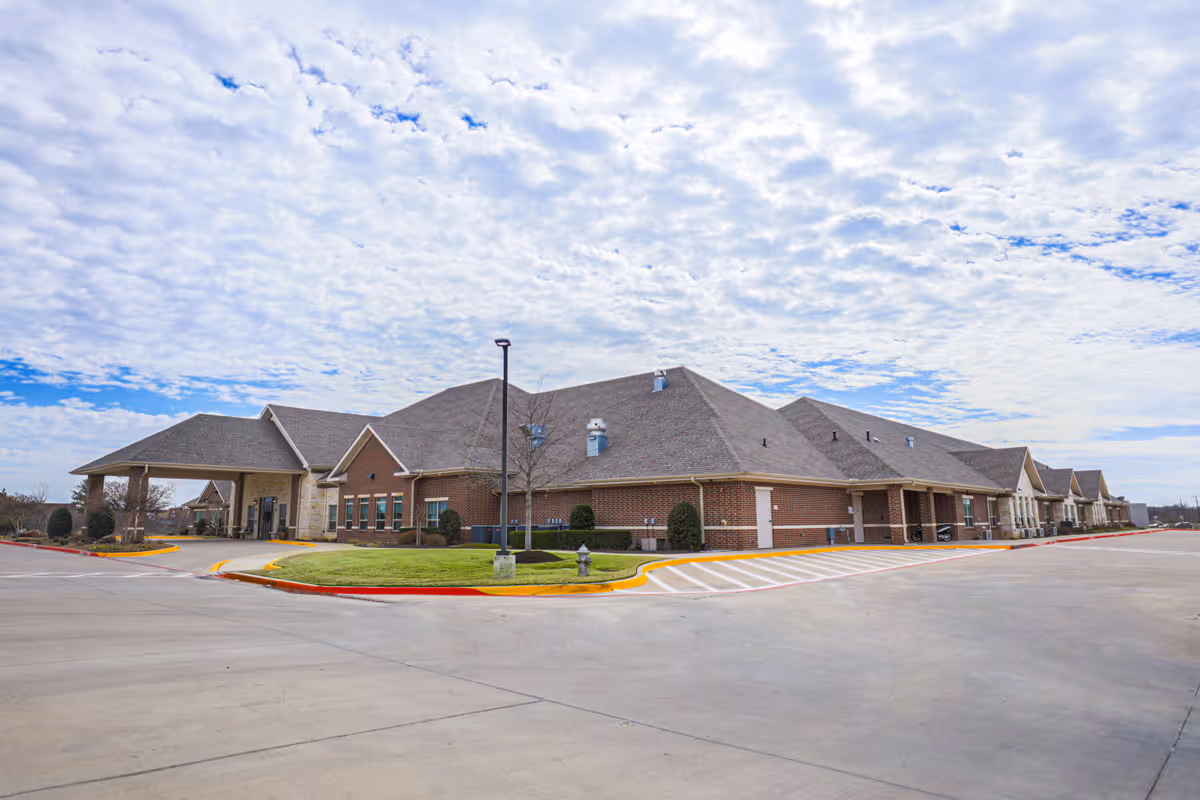 Exterior view of a single-story brick building with a large covered entrance, surrounded by a paved driveway and parking area under a partly cloudy sky.