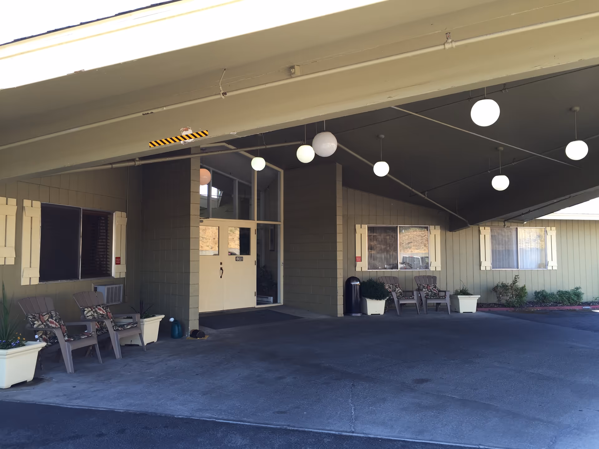 Covered entrance area of a building with beige walls and white double doors. There are several chairs with floral cushions and large white planters with plants on either side of the entrance. Round white hanging lights are suspended from the ceiling.