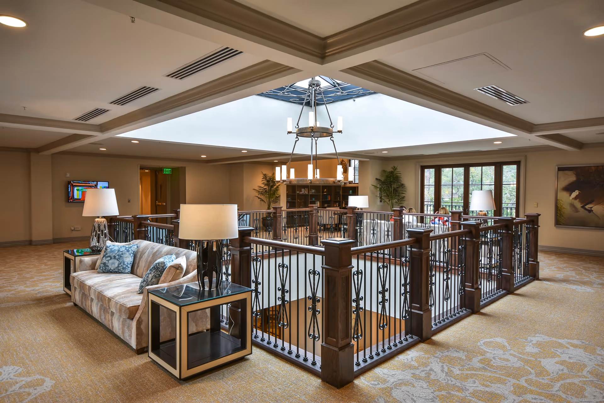 A spacious and well-lit interior common area with a beige carpet and a wooden railing surrounding an open stairwell. There is a beige sofa with patterned cushions and two side tables with lamps. The ceiling features a large skylight with a chandelier hanging in the center. Large windows and plants add to the welcoming atmosphere.