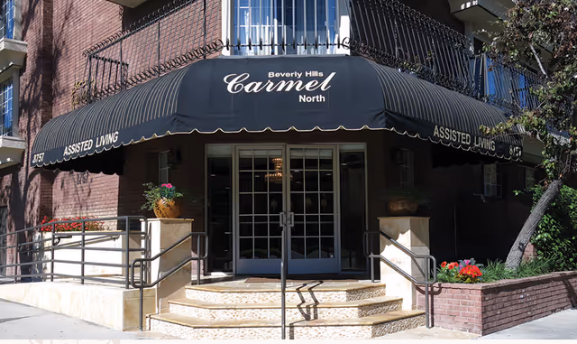 Entrance to a brick building with a black awning that reads 'Beverly Hills Carmel North' and 'Assisted Living' on the sides. There are steps and a ramp leading up to double glass doors, with potted plants and flowers on either side of the entrance.