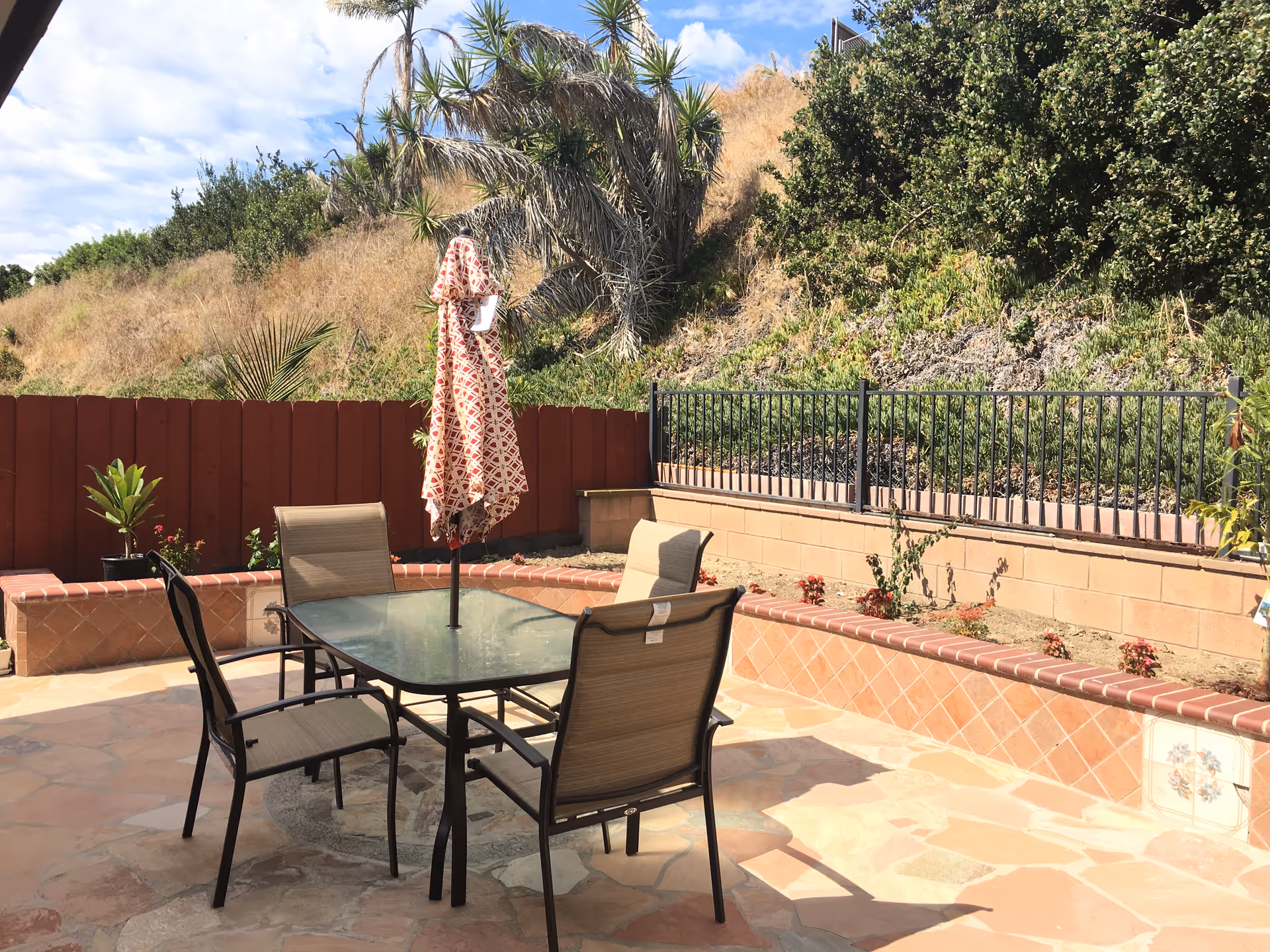 Outdoor patio area with a glass-top table and four chairs. A closed patterned umbrella is positioned in the center of the table. The patio is surrounded by a low brick wall with plants and greenery on the hillside behind it under a partly cloudy sky.