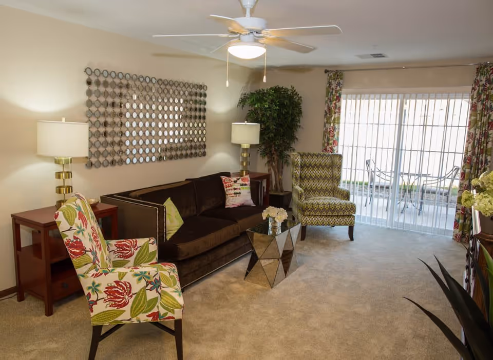 A cozy living room with a dark brown sofa adorned with colorful pillows, two patterned armchairs, a glass coffee table with a flower arrangement, two side tables with lamps, a large decorative mirror on the wall, a ceiling fan with light, and sliding glass doors leading to an outdoor patio with a table and chairs.