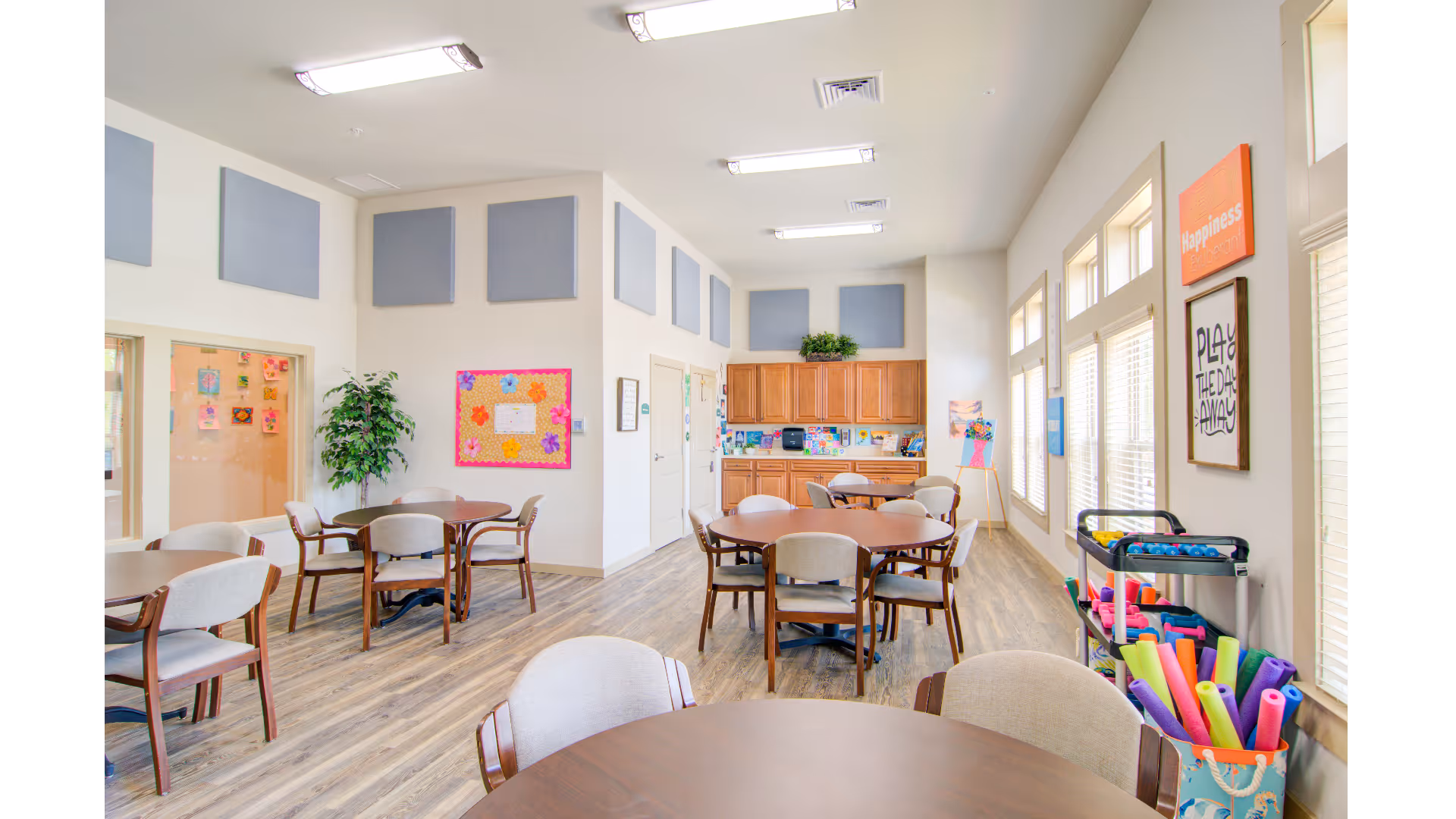 Bright common room with round tables and chairs, cabinets and bulletin boards, and a cart of colorful craft supplies by the windows.