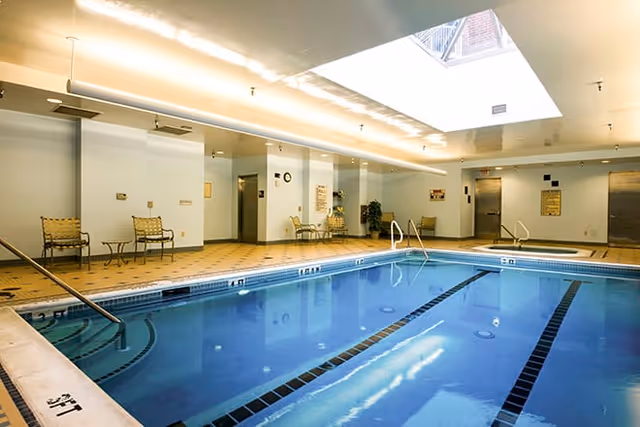 Indoor swimming pool area with clear blue water, surrounded by a tiled deck. There are several chairs and small tables along the walls, with a skylight above providing natural light. The room has a clean, well-lit atmosphere with white walls and ceiling.