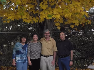 Four adults standing outdoors in front of a tree with yellow leaves and a chain-link fence behind them. They are smiling and posing for the photo.