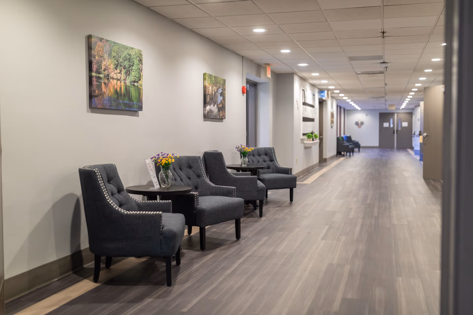 A long, well-lit hallway with gray flooring and white walls. Along one side, there are several dark gray upholstered chairs with nailhead trim, each paired with a small round table holding a vase of flowers. The walls are decorated with framed nature photographs. The hallway extends into the distance with more seating and doors visible.
