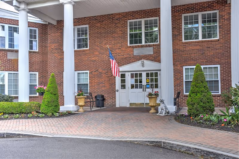 Entrance of a brick building with white columns and multiple windows. There is an American flag mounted near the door, two large planters with flowers, a small statue of a dog, and neatly trimmed bushes and flower beds on either side of the entrance.