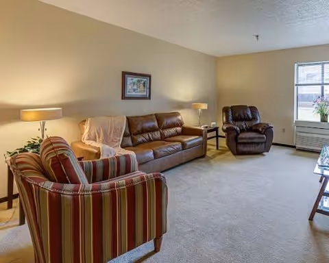 A bright living room with a brown leather sofa, striped armchair, recliner, side tables, lamps, and a window.