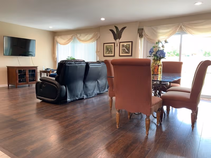 A bright living and dining area with wooden flooring. The living room features a black leather recliner sofa facing a wall-mounted flat-screen TV above a wooden cabinet. The dining area has a glass-top table surrounded by six upholstered chairs in a reddish-brown color. Large windows with sheer curtains allow natural light to fill the space. Decorative framed artwork and a vase with flowers are visible on the walls and table.