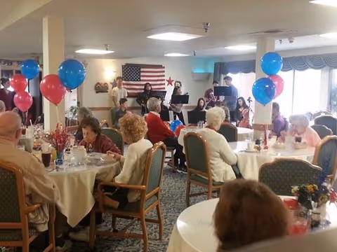 A group of elderly people seated at round tables decorated with red, white, and blue balloons in a dining room. In the background, a small musical group is performing in front of an American flag on the wall. The room has large windows with blue curtains and is well-lit with ceiling lights.