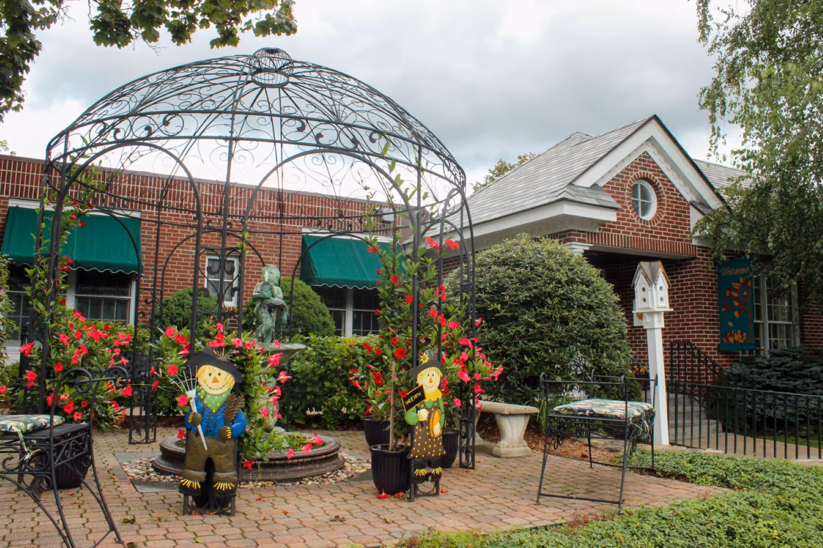 Outdoor garden area at Blaire House of Milford featuring a decorative metal gazebo with climbing plants and flowers, two scarecrow decorations, a small fountain with a statue, brick building with green awnings, and surrounding greenery.
