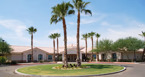 Exterior view of a single-story building with a circular driveway and a landscaped roundabout featuring several tall palm trees and green grass under a blue sky with some clouds.