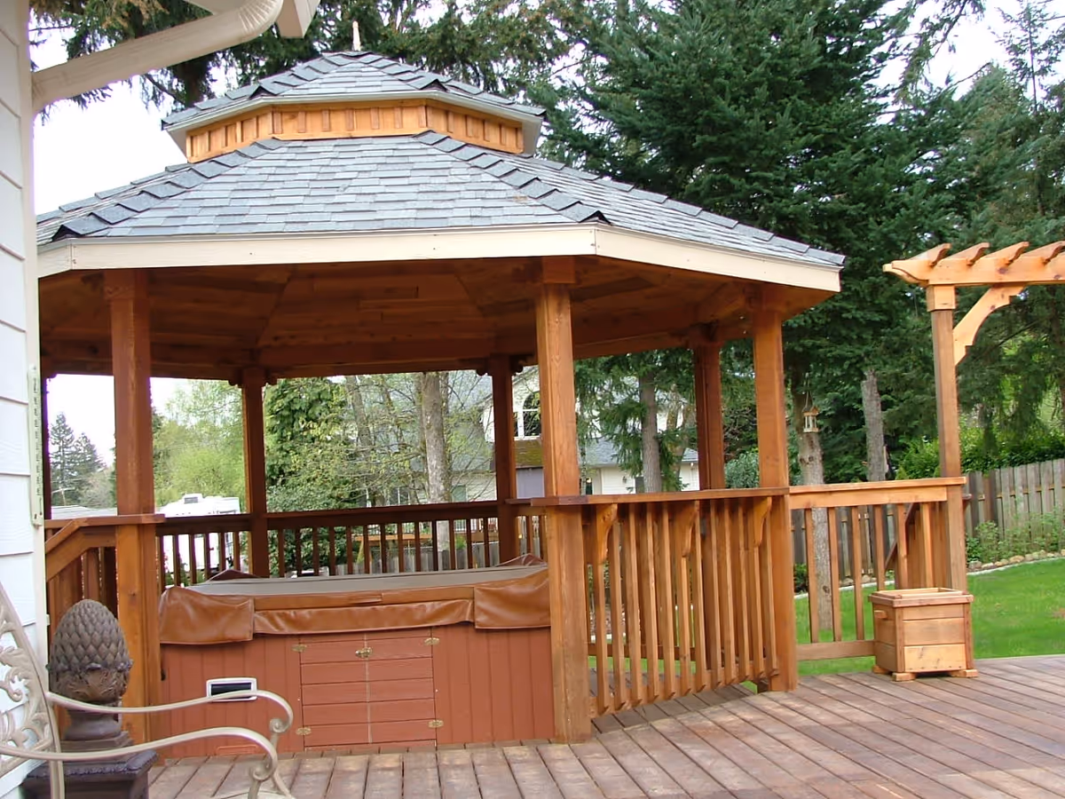 A wooden gazebo with a shingled roof situated on a wooden deck. The gazebo has railings and contains a covered hot tub. Surrounding the deck are green trees and a wooden fence in the background.