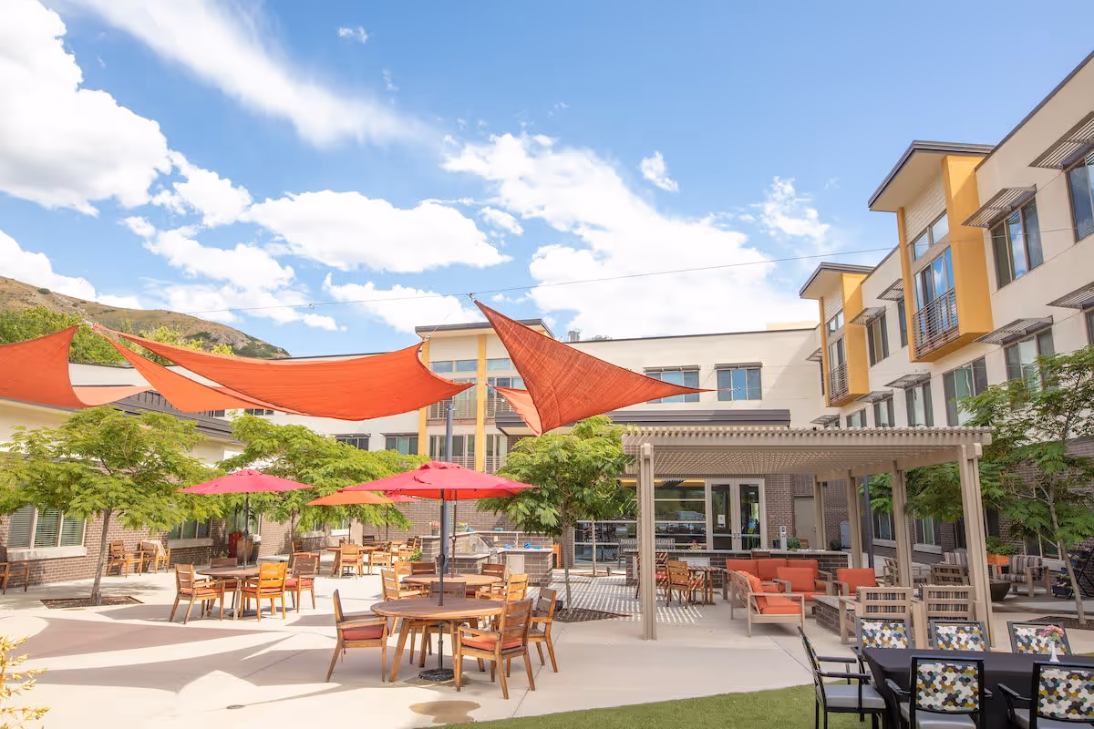 Sunlit outdoor courtyard with tables, chairs, red umbrellas and shade sails in front of a multi-story senior living building.