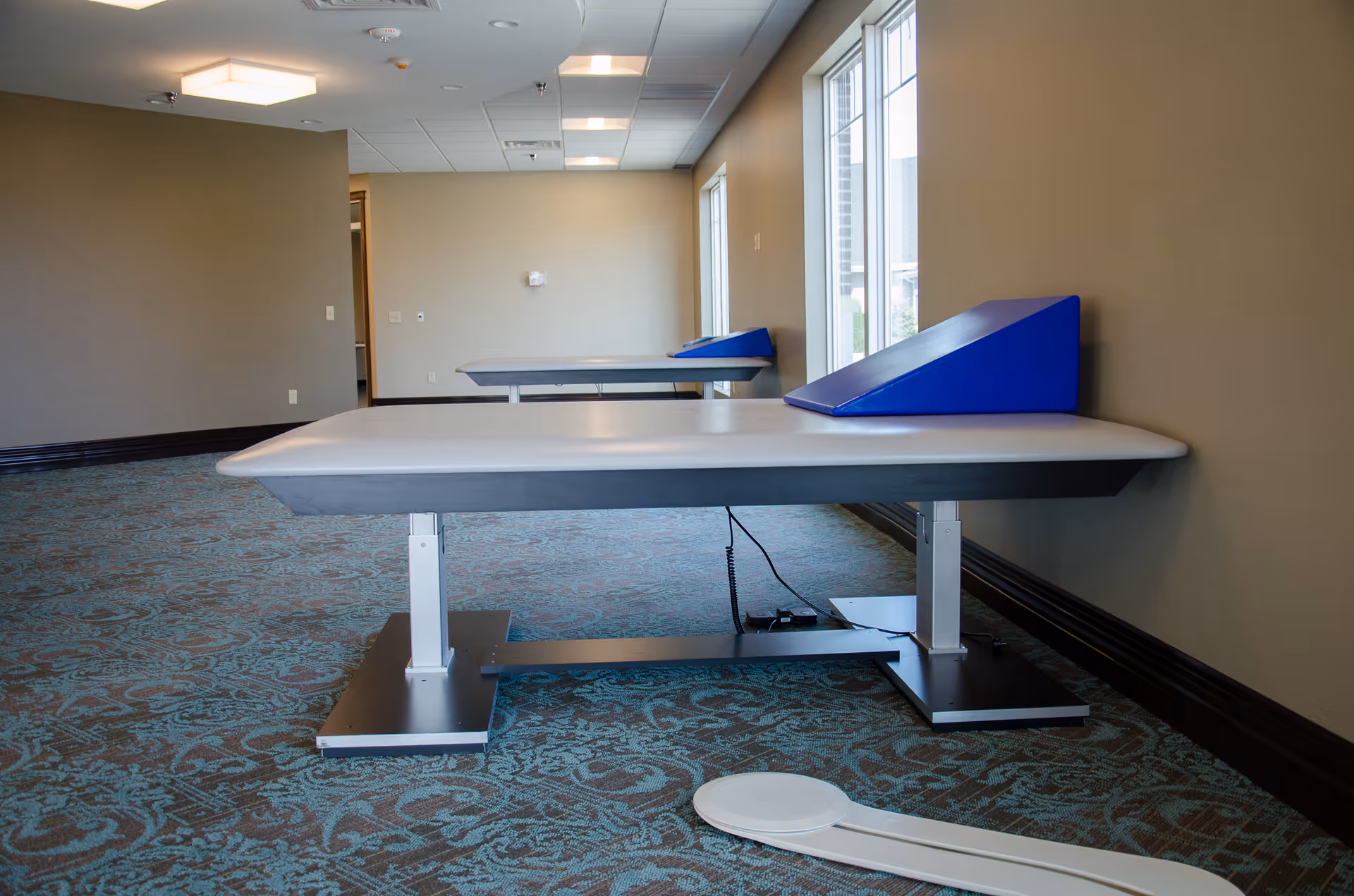 A room with two medical examination or therapy tables with blue wedge cushions against the wall, patterned carpet flooring, beige walls, and large windows letting in natural light.