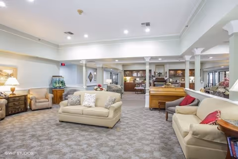 A spacious and well-lit living room area in a senior living facility featuring beige sofas with decorative pillows, armchairs, a wooden piano, a fish tank, and built-in shelves with various decorative items. The room has carpeted flooring, white walls, and recessed lighting in the ceiling.
