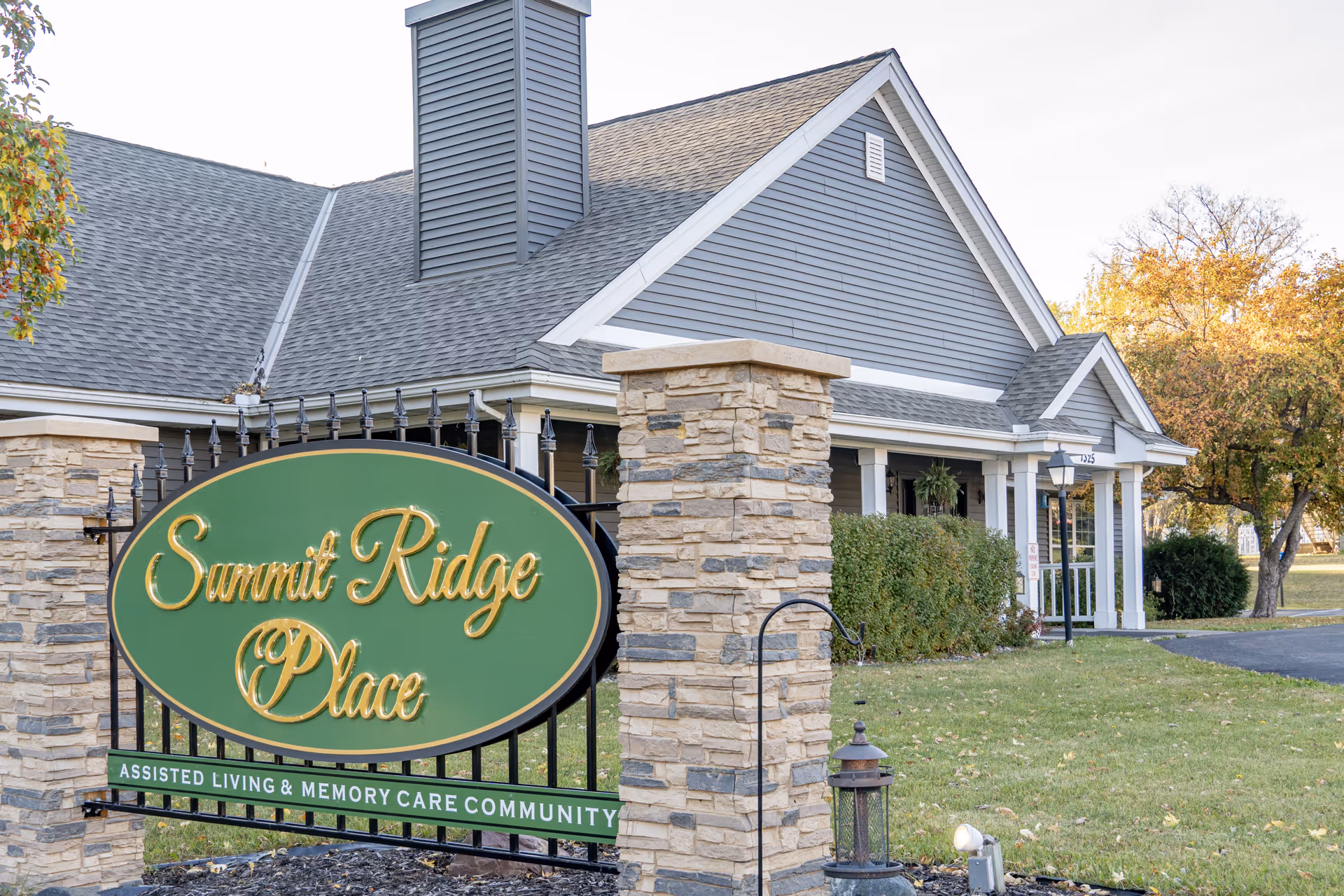 Exterior view of Summit Ridge Place, an assisted living and memory care community, showing a stone and metal sign with the facility name in front of a gray building with white trim and a porch, surrounded by green grass and trees with autumn foliage.