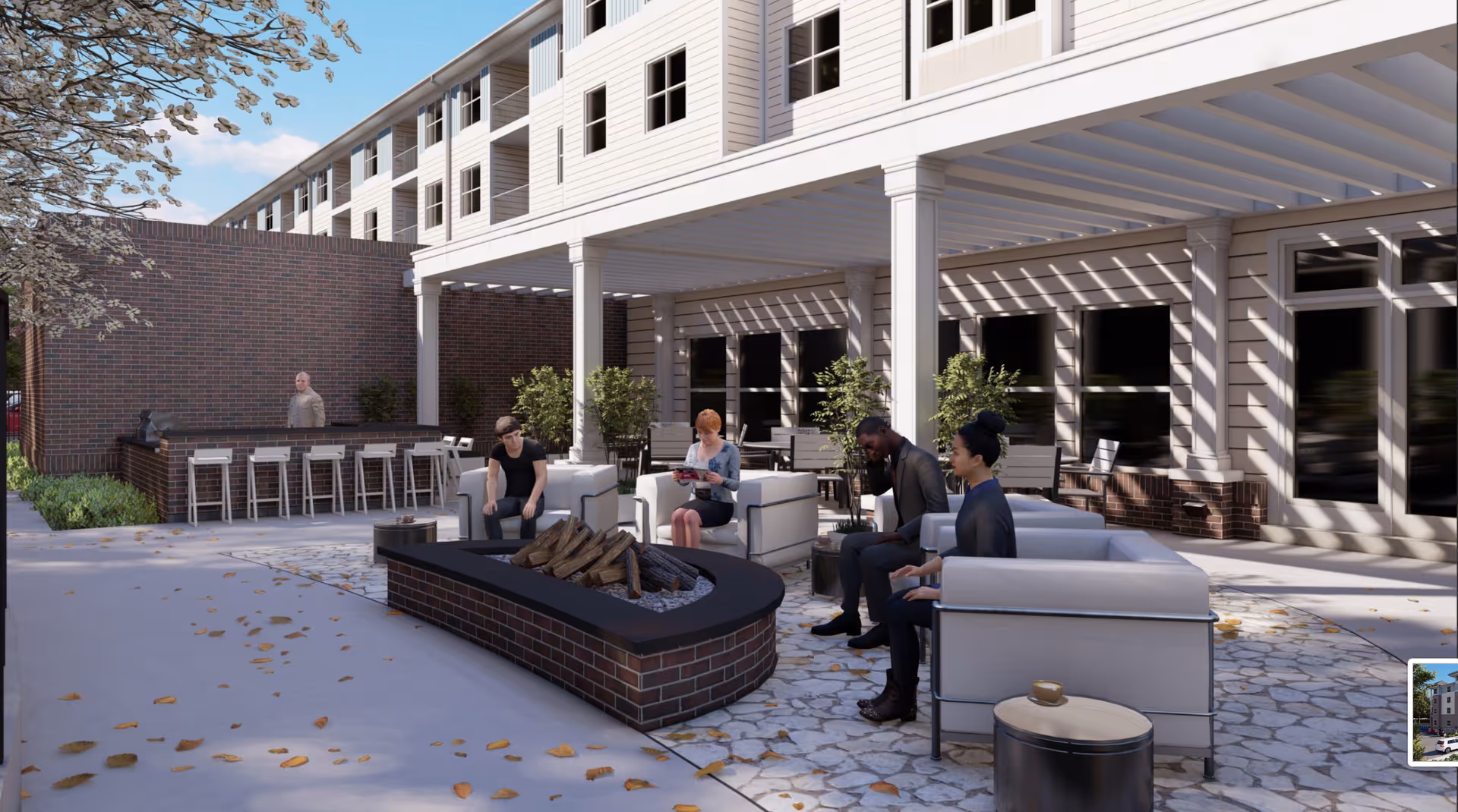Outdoor patio area at Acclaim at Cary Pointe with modern seating around a brick fire pit. Four people are seated on white cushioned chairs, and one person is standing behind a bar counter. The building exterior features white siding and large windows, with a pergola casting shadows on the patio floor scattered with fallen leaves.