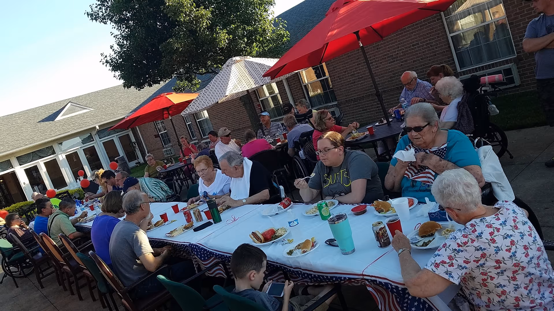 A group of elderly people and some younger individuals sitting around long tables outdoors, enjoying a meal together under large umbrellas. The setting appears to be a courtyard of a brick building with windows and a green lawn.