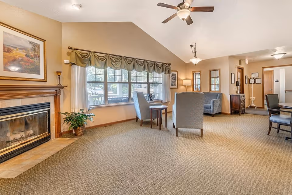 A cozy living room area in a senior living facility with a fireplace on the left, a large window with green valance curtains, two armchairs and a small table in the center, a sofa in the background, ceiling fan, and warm lighting.
